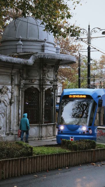 A historic ornate stone kiosk with intricate carvings and a domed roof is situated along a street near a tram stop in Addiscombe. The kiosk features decorative columns and lattice windows, with a weat